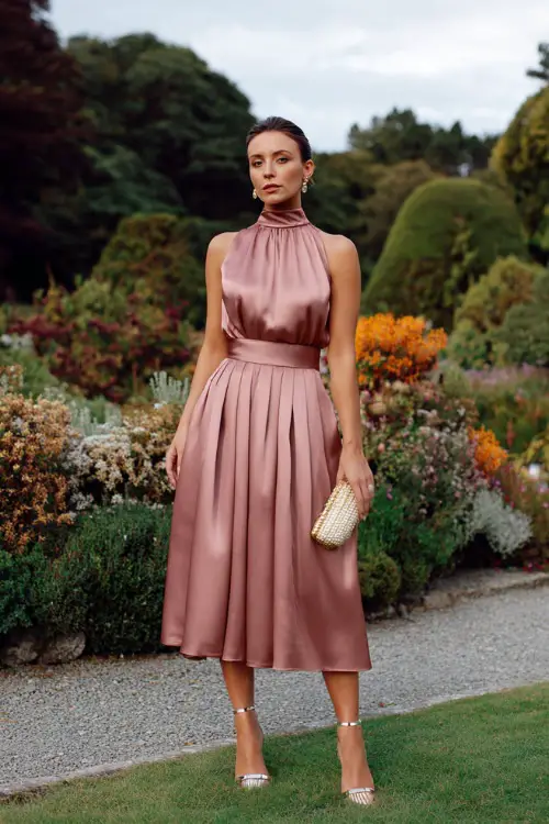 A woman wears a blush satin high-neck midi dress with a pleated skirt, metallic sandals, and a pearl-embellished clutch in an autumn garden venue 