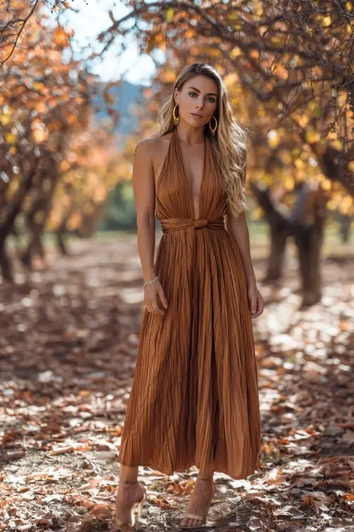 A woman wears a bronze pleated maxi dress with halter neckline, styled with hoop earrings and strappy heels, posing in an orchard with autumn leaves scattered across the ground