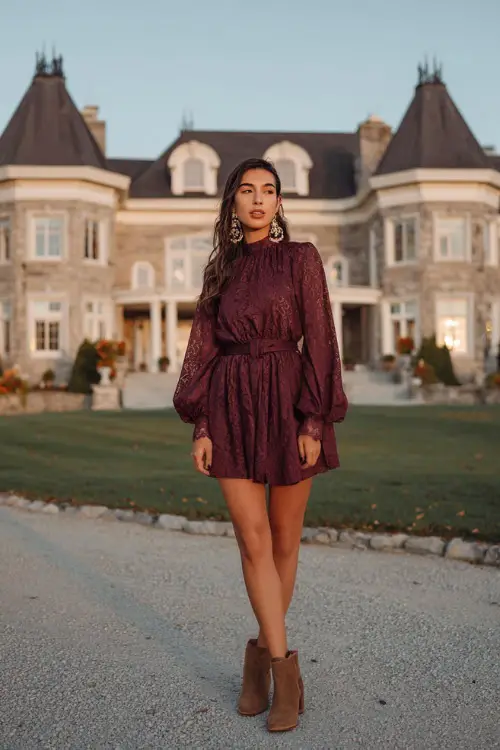 A woman wears a burgundy A-line dress with a cinched waist and lace overlay, styled with brown ankle boots and statement earrings, posing near a grand estate decorated for fall weddings 