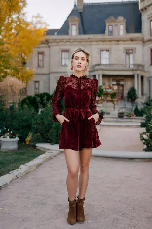 A woman wears a burgundy A-line dress with a cinched waist and lace overlay, styled with brown ankle boots and statement earrings, posing near a grand estate decorated for fall weddings