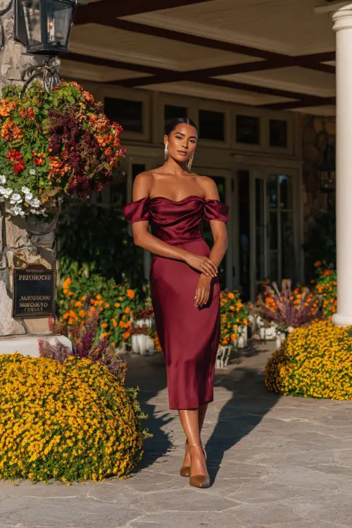 A woman wears a burgundy satin midi dress with an off-the-shoulder neckline, styled with gold heels and pearl earrings 