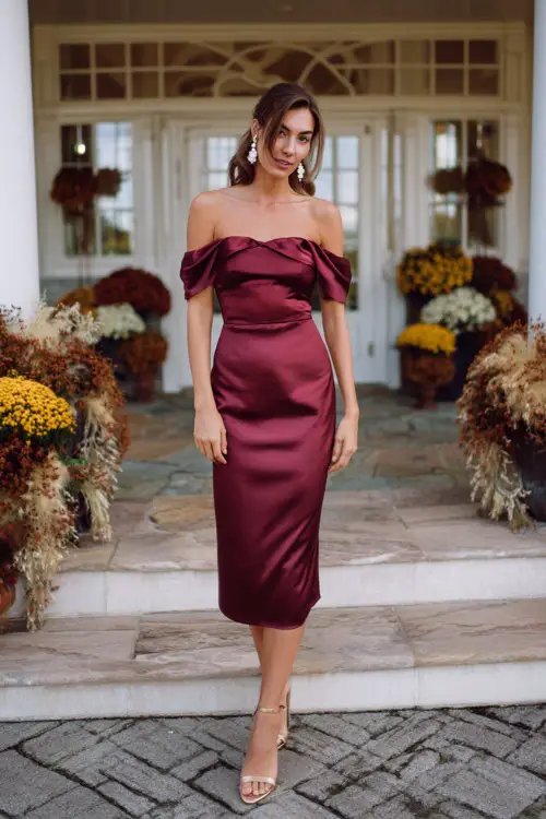 A woman wears a burgundy satin midi dress with an off-the-shoulder neckline, styled with gold heels and pearl earrings