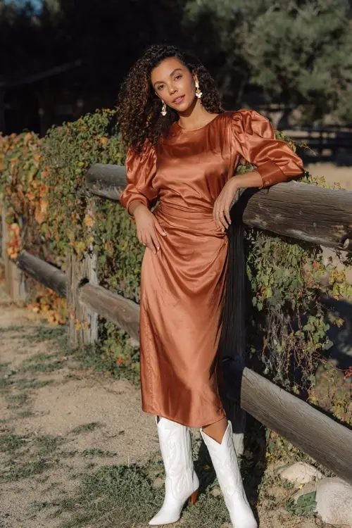 A woman wears a copper satin midi dress with puff sleeves, accessorized with white cowboy boots and statement earrings, standing near a rustic wooden fence lined with fall ivy