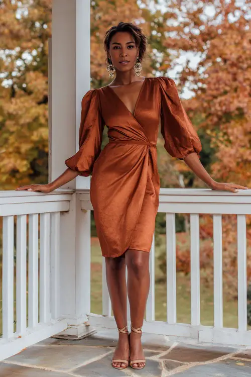 A woman wears a copper satin wrap dress with a V neckline and puff sleeves, styled with statement earrings and strappy heels, standing on a veranda with fall foliage in the background