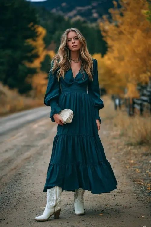 A woman wears a dark teal chiffon maxi dress with tiered skirt, paired with white cowboy boots and a matching clutch, standing along a country road lined with October foliage 