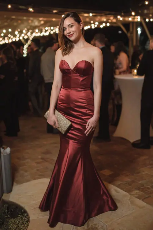 A woman wears a deep red satin mermaid gown with a sweetheart neckline, styled with a matching clutch and metallic heels, standing beneath twinkling lights at a fall wedding reception 