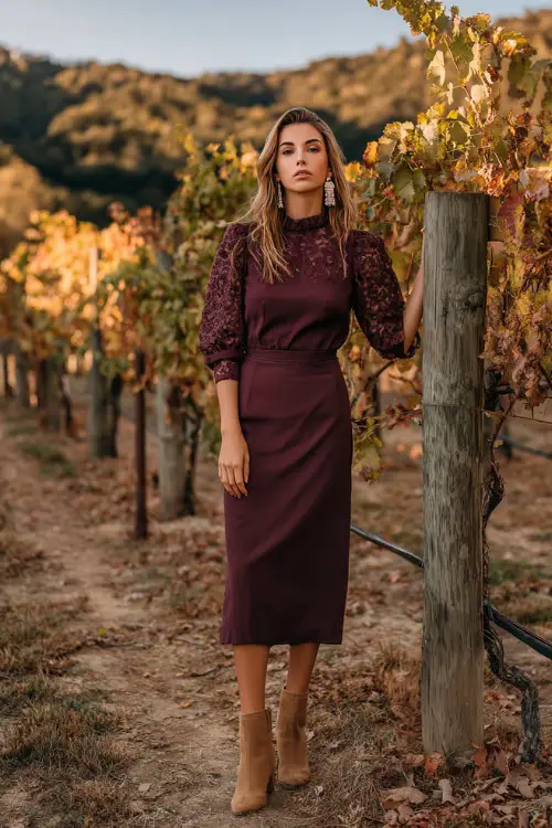 A woman wears a maroon midi dress with lace sleeves, styled with tan block-heeled boots and pearl earrings, standing at a vineyard terrace with fall vines around her 