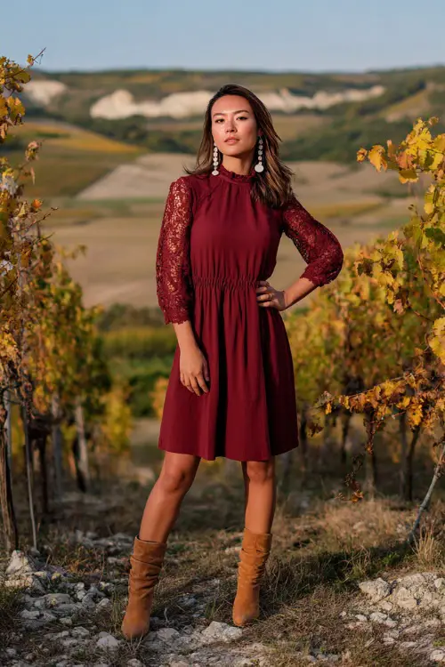 A woman wears a maroon midi dress with lace sleeves, styled with tan block-heeled boots and pearl earrings, standing at a vineyard terrace with fall vines around her