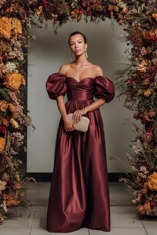 A woman wears a maroon satin gown with puff sleeves and floor-sweeping skirt, styled with a jeweled clutch and earrings, standing near a grand wedding arch covered in autumn florals 