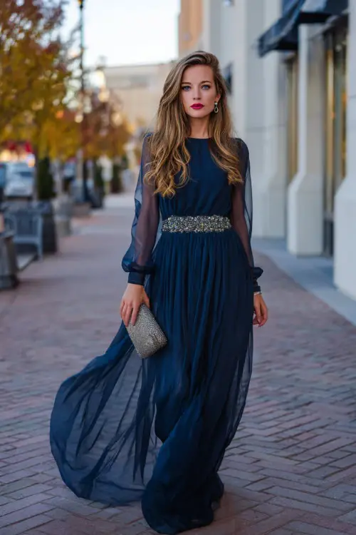 A woman wears a midnight blue chiffon ball gown with sheer long sleeves and a jeweled belt, styled with silver heels and a metallic clutch for a formal autumn celebration 