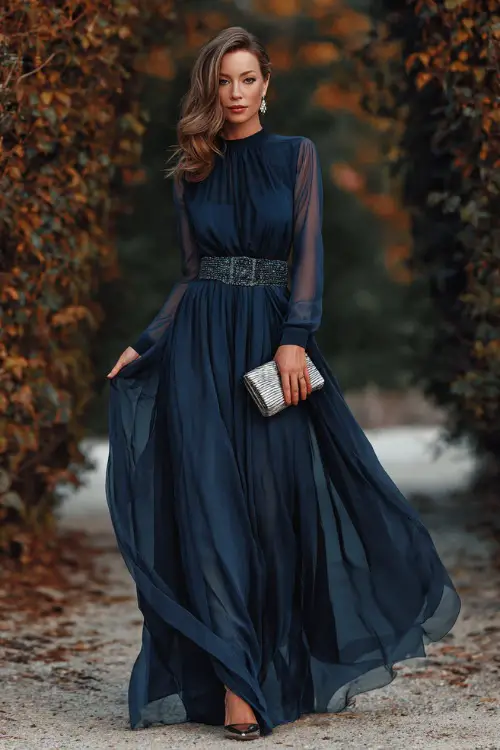 A woman wears a midnight blue chiffon ball gown with sheer long sleeves and a jeweled belt, styled with silver heels and a metallic clutch for a formal autumn celebration