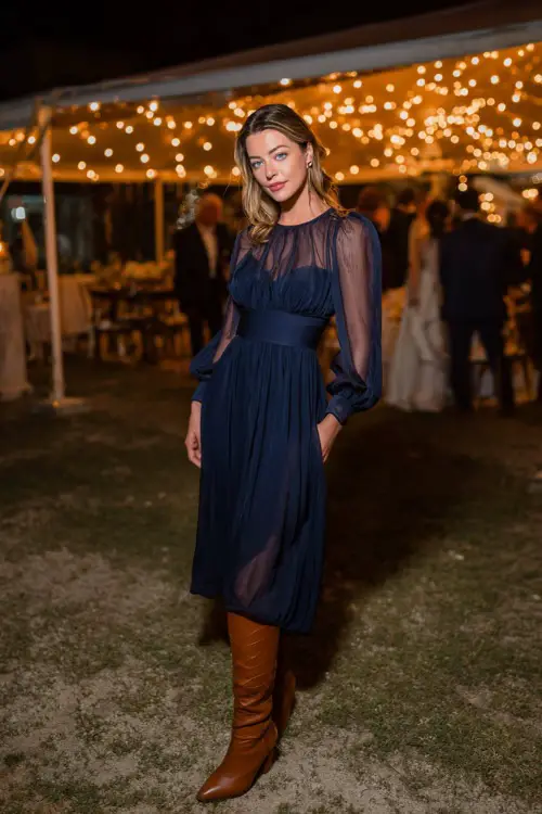A woman wears a navy chiffon midi dress with sheer bishop sleeves, accessorized with tall tan leather boots, standing beneath twinkling lights at an outdoor wedding reception 