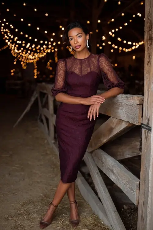 A woman wears a plum lace midi dress with sheer sleeves, accessorized with pearl drop earrings and heels, standing in a barn venue lit with twinkling string lights 
