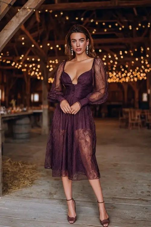 A woman wears a plum lace midi dress with sheer sleeves, accessorized with pearl drop earrings and heels, standing in a barn venue lit with twinkling string lights