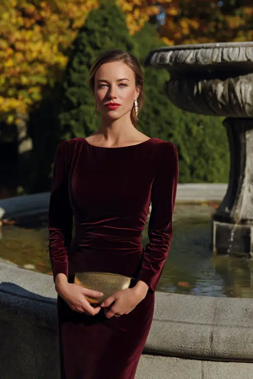 A woman wears a simple burgundy velvet column gown with long sleeves, accessorized with a gold clutch and earrings 