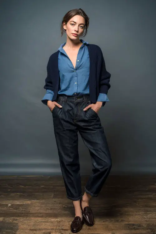 Elegant woman in high-rise dark denim paired with a button-down blue shirt and navy pullover sweater draped over the shoulders, styled with classic loafers 