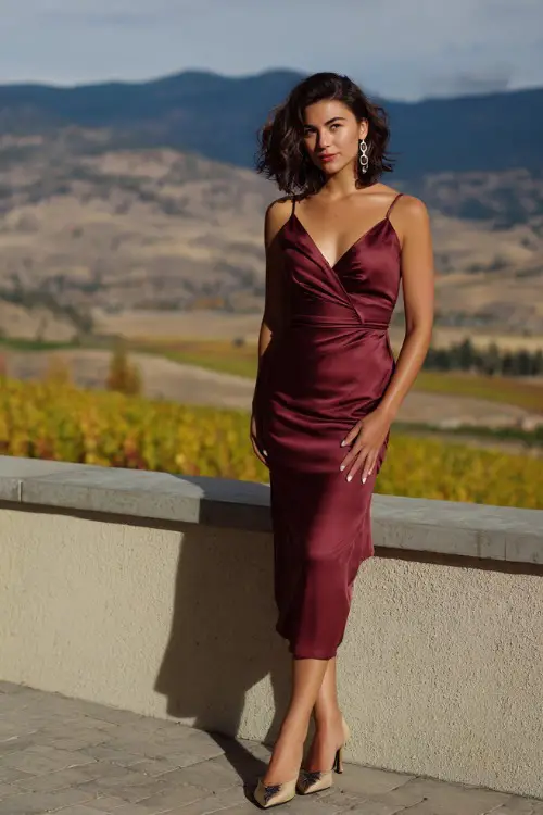 Woman in a burgundy satin midi dress with a fitted bodice and subtle side slit, styled with drop earrings and kitten heels