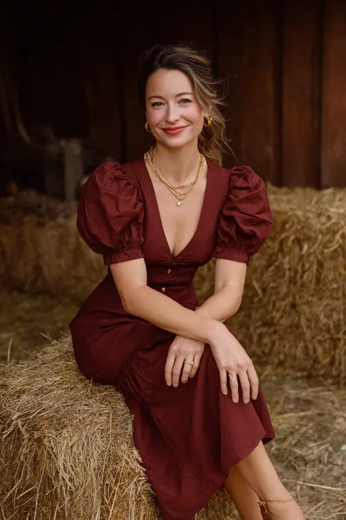 Woman in a burgundy smocked midi dress with puff sleeves, styled with loafers and a thin gold necklace, sitting on a hay bale at a rustic October wedding