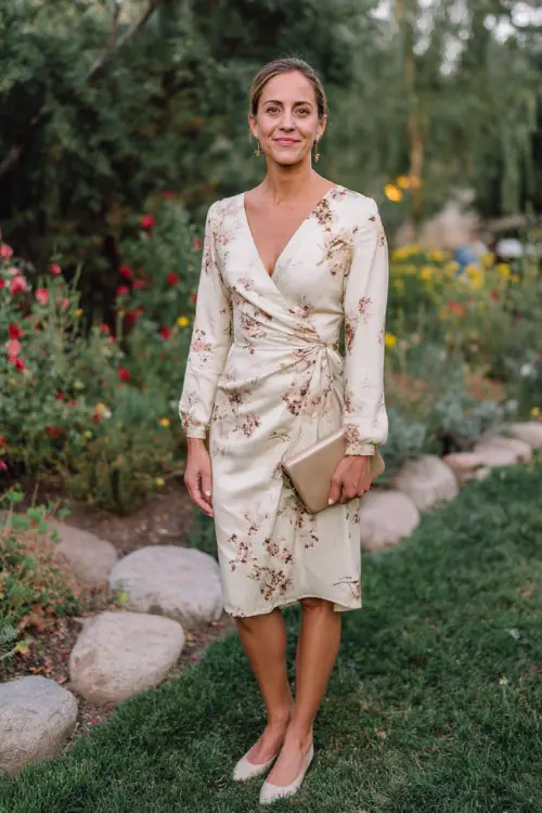 Woman in a cream floral-print wrap midi dress with long sleeves, paired with ballet flats, holding a small clutch at a laid-back garden wedding in October 