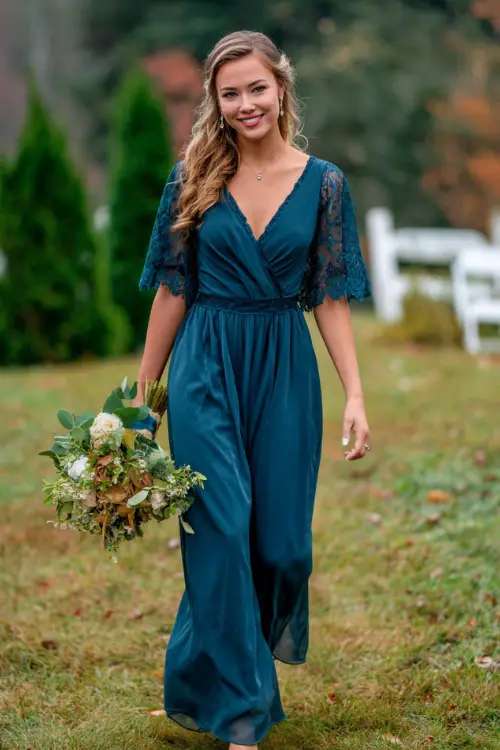 Woman in a dark teal A-line dress with lace accents and flutter sleeves, holding a bouquet while walking on a grassy outdoor aisle in fall