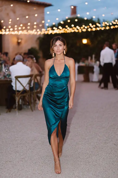 Woman in a dark teal satin midi dress with side slit and halter neckline, paired with gold accessories and strappy heels, walking past string lights at a semi-formal October evening wedding 