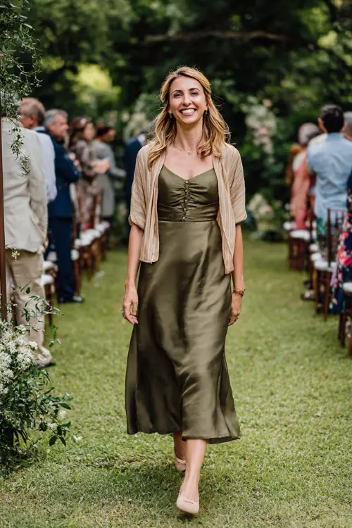 Woman in a deep olive green A-line midi dress with short sleeves and a flowy skirt, styled with ballet flats and a lightweight cardigan, walking along a leafy outdoor wedding aisle 