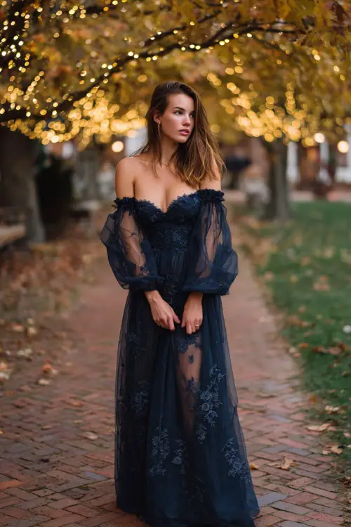 Woman in a floor-length navy gown with sheer sleeves and floral embroidery, standing under an autumn tree canopy decorated with fairy lights 