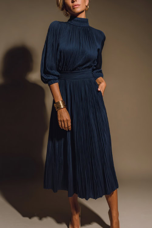 Woman in a high-neck navy midi dress with pleated hem and cuffed sleeves, styled with a gold bracelet and low heels, soft-lit background