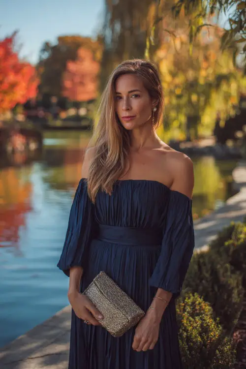 Woman in a midnight blue off-the-shoulder dress with a pleated skirt, accessorized with a metallic clutch 