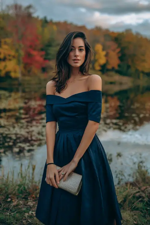 Woman in a midnight blue off-the-shoulder dress with a pleated skirt, accessorized with a metallic clutch 