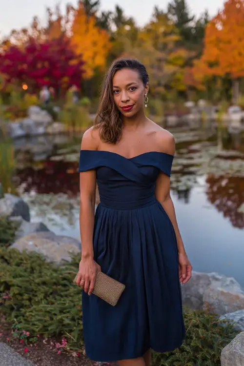 Woman in a midnight blue off-the-shoulder dress with a pleated skirt, accessorized with a metallic clutch 