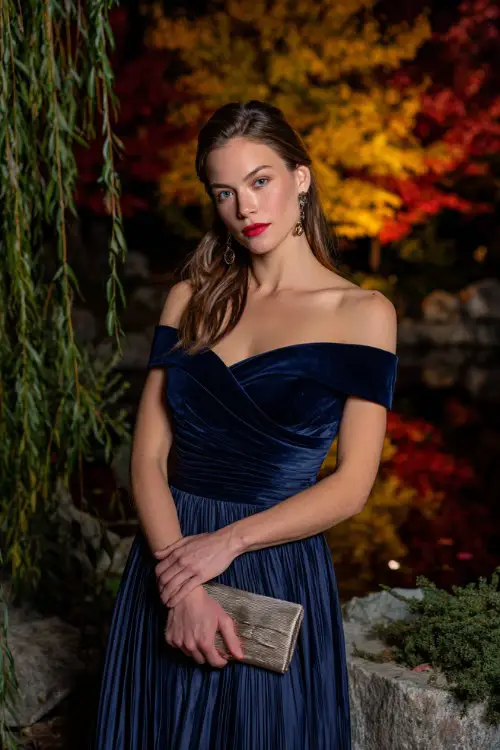 Woman in a midnight blue off-the-shoulder dress with a pleated skirt, accessorized with a metallic clutch