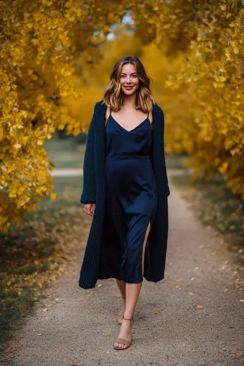 Woman in a navy cowl-neck slip dress with a long cardigan layered on top, styled with heeled mules, standing in a golden-leaf park wedding venue