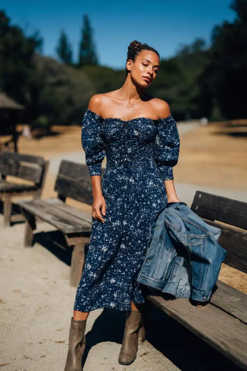Woman in a navy floral-print midi dress with puff sleeves and a smocked bodice, holding a denim jacket and wearing low-heeled boots, standing by wooden wedding benches in a park