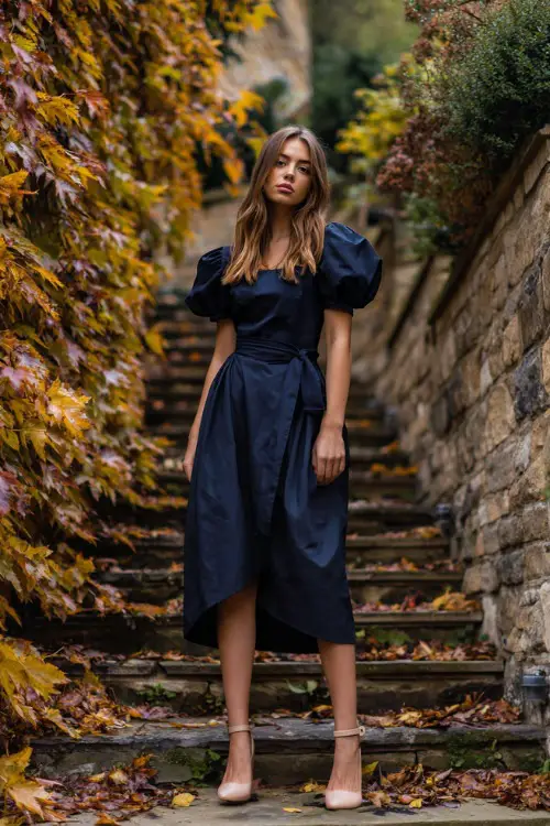 Woman in a navy high-low hem midi dress with puff sleeves, styled with block-heeled sandals, standing beside a stone pathway surrounded by autumn leaves