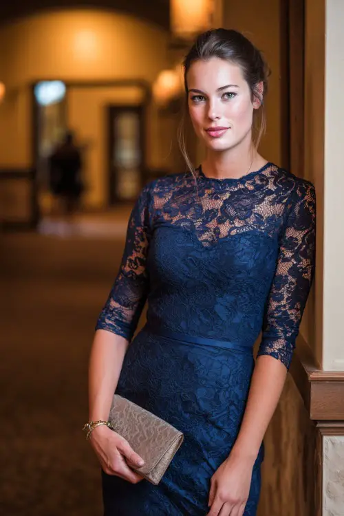 Woman in a navy lace-overlay sheath dress with three-quarter sleeves, holding a small clutch, posing inside a warmly lit October wedding hall, full body 