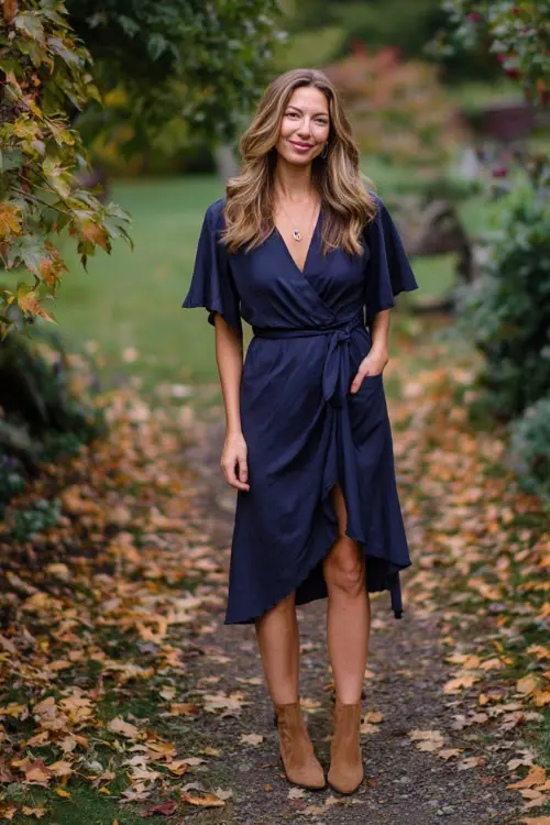 Woman in a navy wrap midi dress with flutter sleeves and a tie waist, paired with tan ankle boots, standing on a leaf-covered garden path at an October wedding 