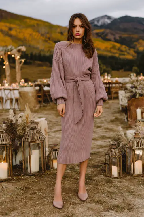 Woman in a soft mauve sweater dress with a belted waist and midi length, paired with heeled mules, standing near a rustic outdoor October wedding setup with lanterns