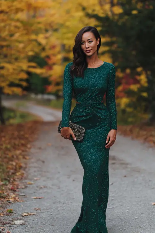 Woman in an emerald green long sleeve fitted gown with subtle sequins, styled with a matching shawl and evening clutch, standing on a fall foliage-lined pathway 