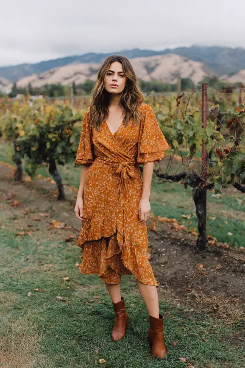 Woman wearing a burnt orange floral midi dress with flutter sleeves, paired with leather ankle boots, standing near a vineyard wedding backdrop in October