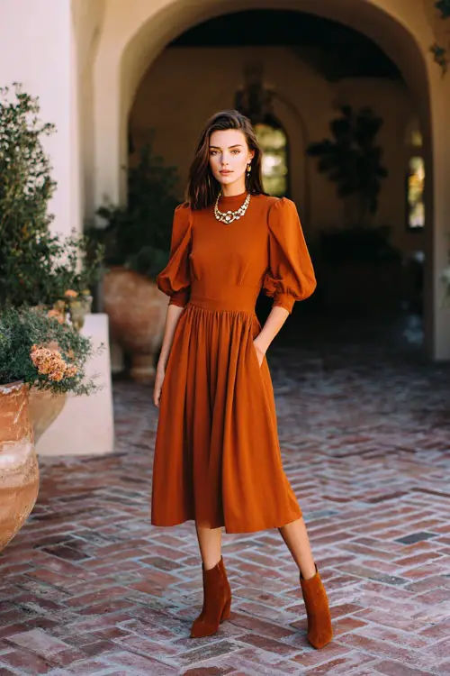 Woman wearing a burnt sienna fit-and-flare midi dress with elbow-length sleeves, styled with suede ankle boots and a statement necklace, standing in a warm-lit October courtyard wedding 