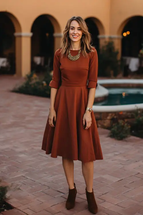 Woman wearing a burnt sienna fit-and-flare midi dress with elbow-length sleeves, styled with suede ankle boots and a statement necklace, standing in a warm-lit October courtyard wedding