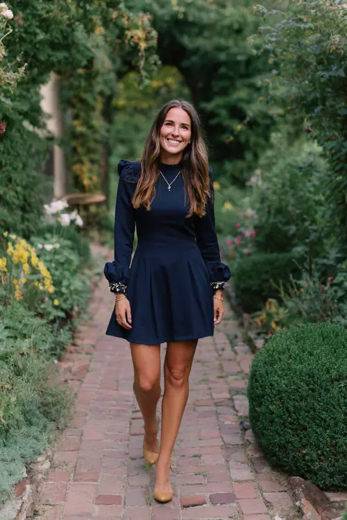 Woman wearing a navy fit-and-flare dress with embroidered cuffs, paired with low block heels and a pendant necklace, walking along a cozy October garden wedding path