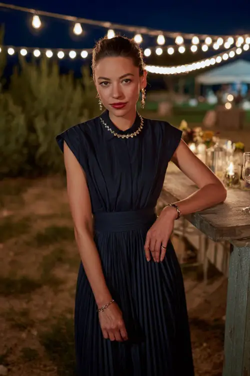 Woman wearing a navy pleated tea-length dress with cap sleeves and silver jewelry, posing by an outdoor reception table under string lights, full body 