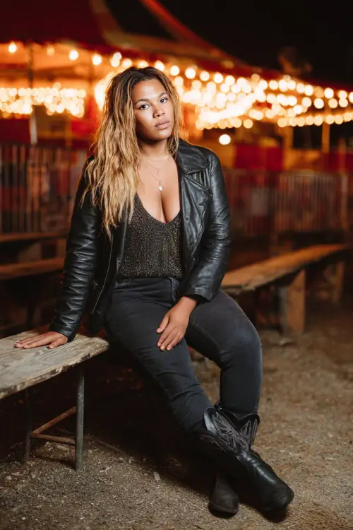 A plus-size woman dressed in a leather jacket layered over a sequin cami, black jeans, and black cowboy boots, posing near a night concert stage with glowing lights 