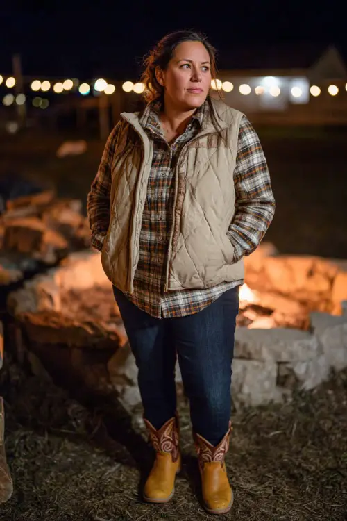 A plus-size woman in a quilted puffer vest layered over a flannel shirt, dark jeans, and tan cowboy boots, standing near a firepit with glowing lights in the distance 