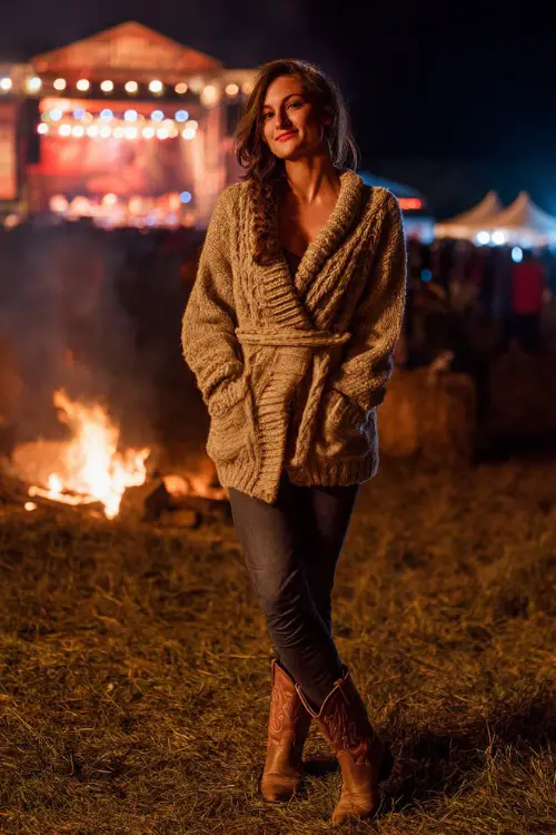 A woman in a chunky knit cardigan belted at the waist, dark skinny jeans, and cowboy boots, standing beside a bonfire with a concert stage glowing in the background 
