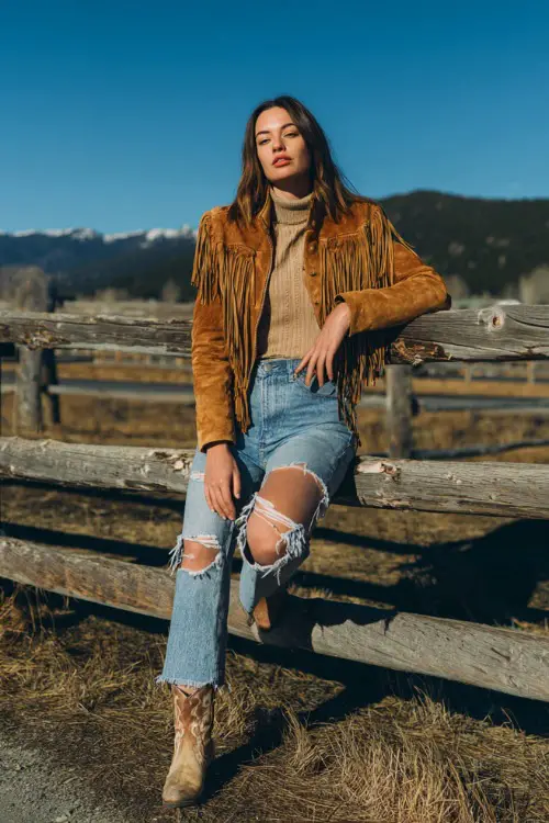 A woman in a suede fringe jacket over a turtleneck top, ripped jeans, and ankle cowboy boots, leaning against a wooden fence at a country concert