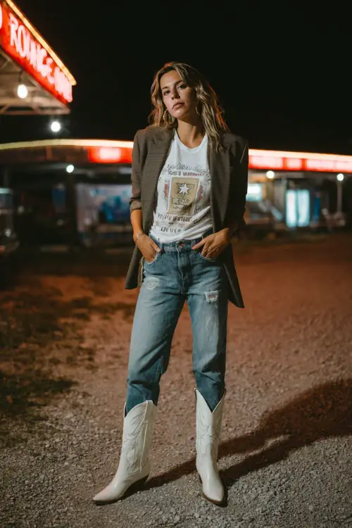A woman in distressed blue jeans, graphic tee, and tailored blazer, paired with pointed white cowboy boots, posing confidently near neon lights at a night country concert
