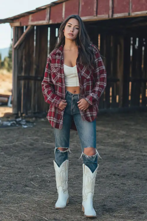 A woman in ripped skinny jeans, oversized plaid flannel, and sleek white cowboy boots, standing in front of a rustic barn at a fall country concert